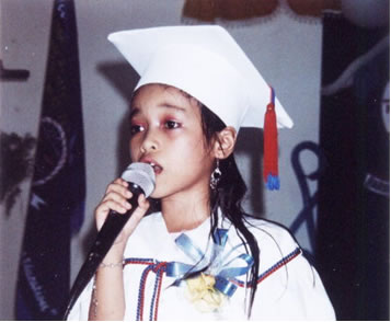Girl in white graduation cap and robe singing at microphone.