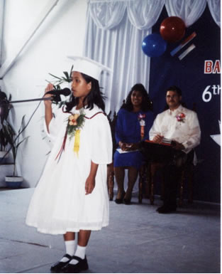 Young girl in white graduation robe and mortarboard speaking into microphone from BLC stage.